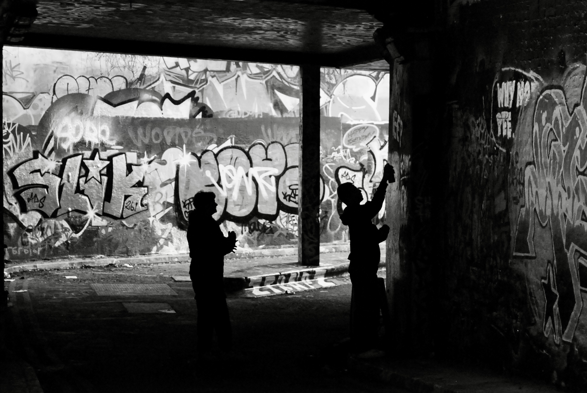 To young men in silhouette spray paint a message on the wall of a tunnel
