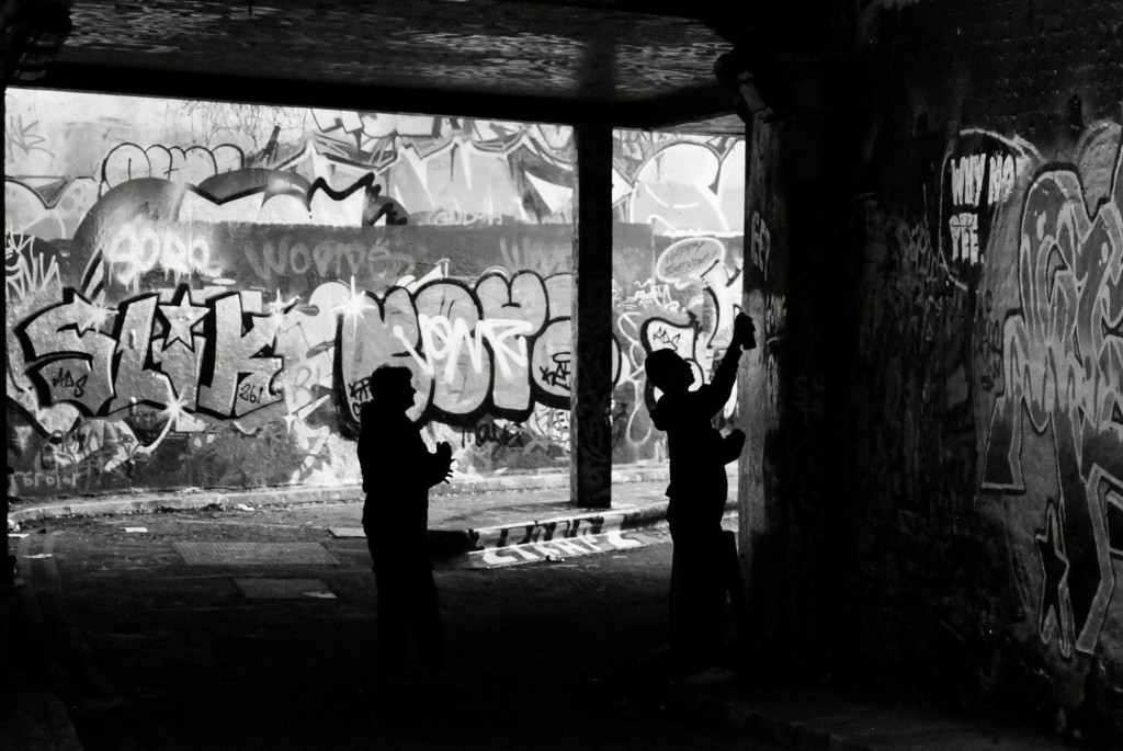 To young men in silhouette spray paint a message on the wall of a tunnel